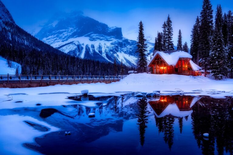 snowy lakeside cabin below the mountains illuminated from the inside at dusk