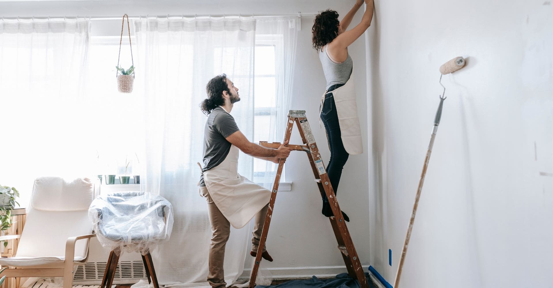 a man holding a ladder while I woman stands on top to paint a wall