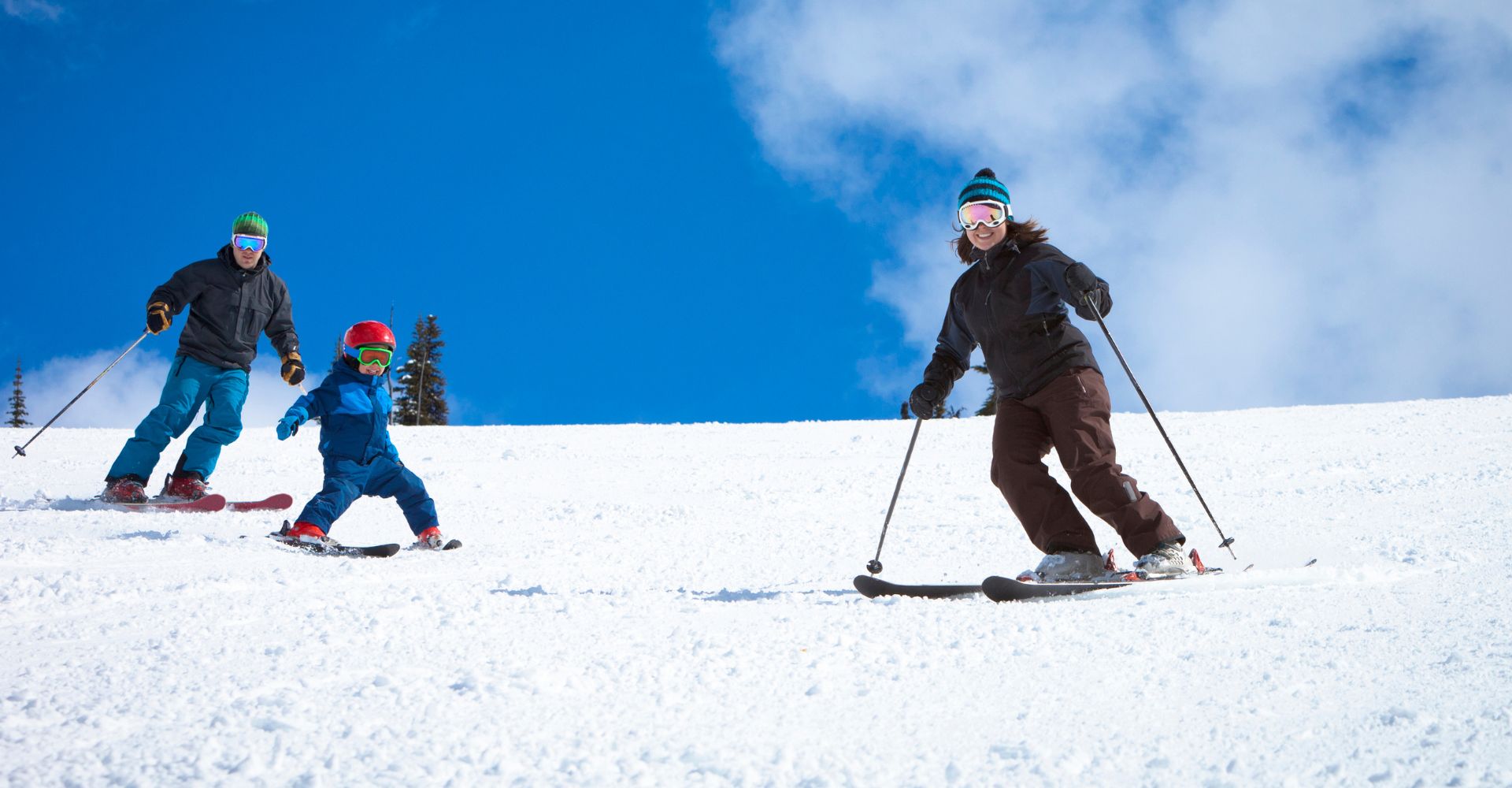 A family enjoys skiing together on a snowy hill, surrounded by trees and a clear blue sky