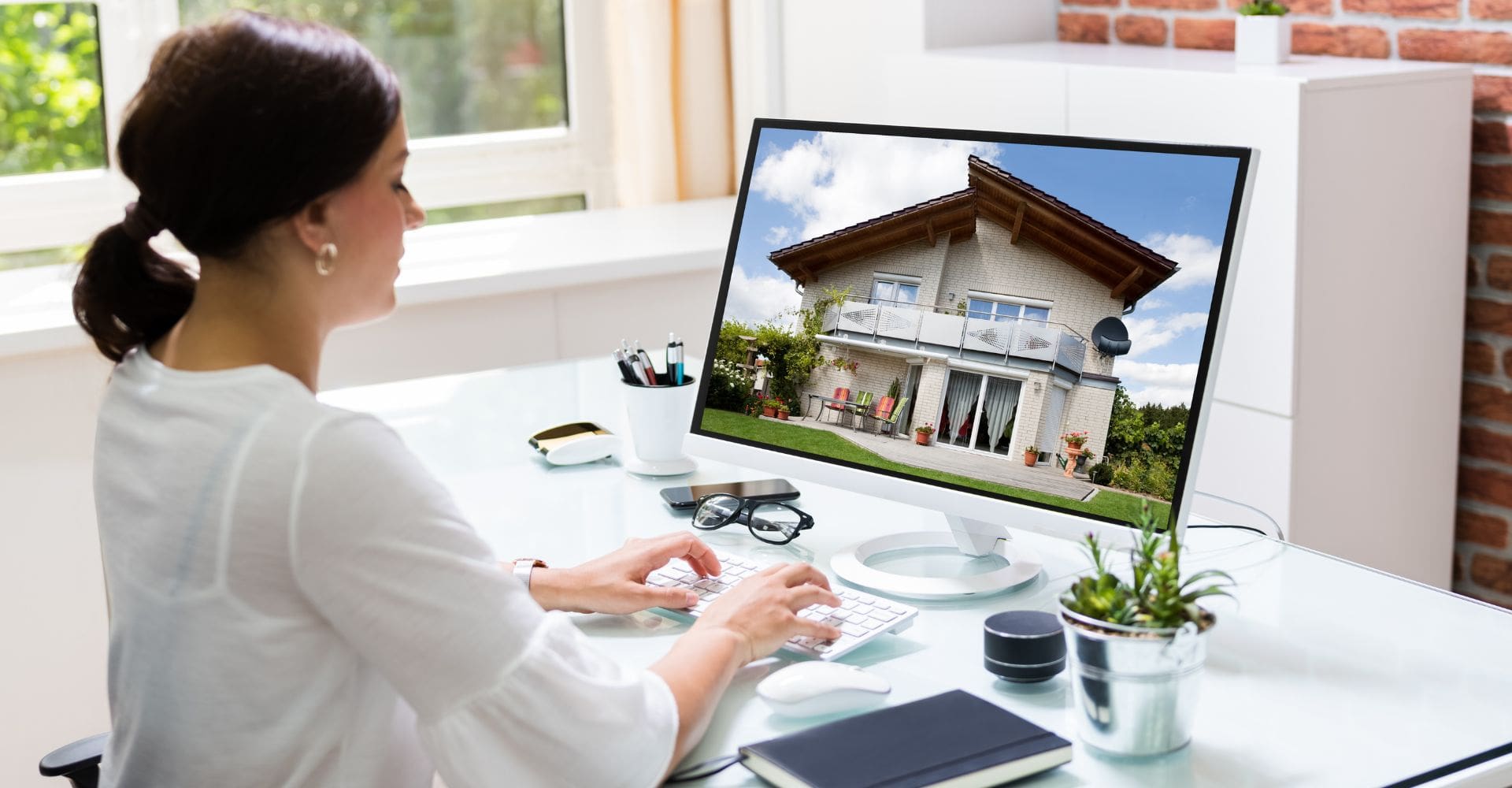 A woman sits at a desk, focused on a computer screen displaying an image of a house
