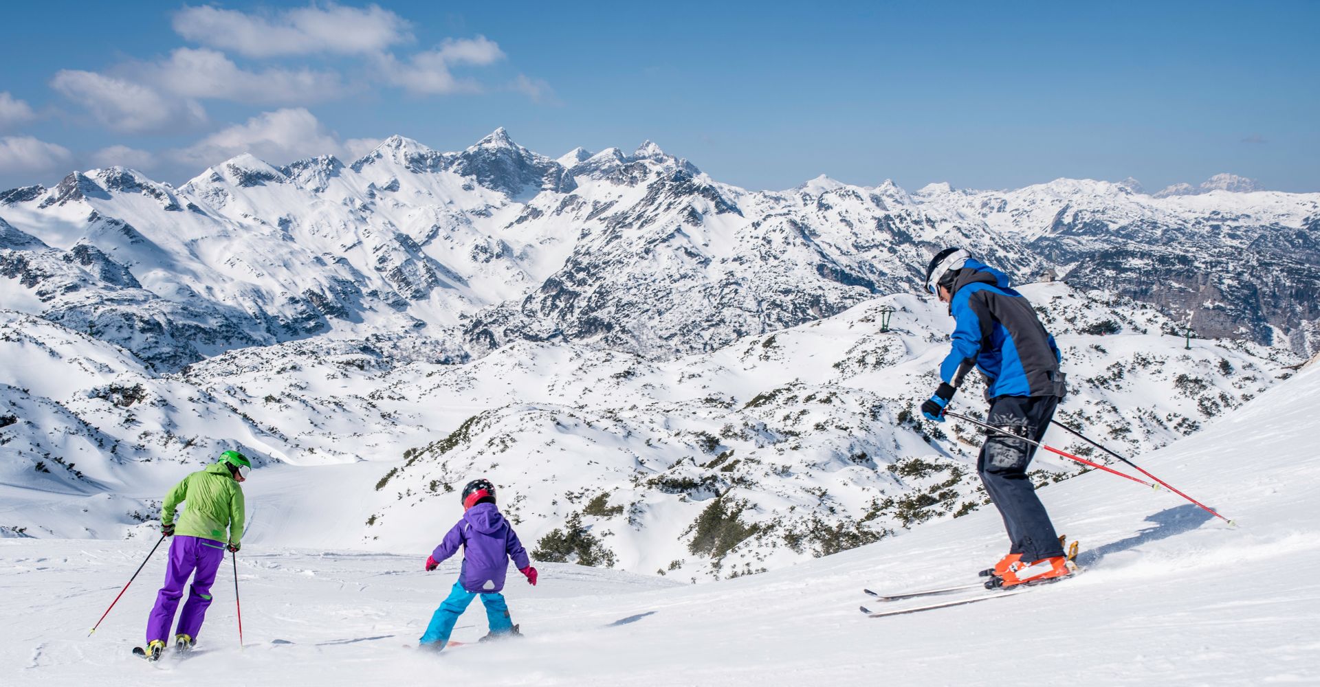 A family of three skiers stand on a snowy mountain, surrounded by a pristine winter landscape under a clear blue sky