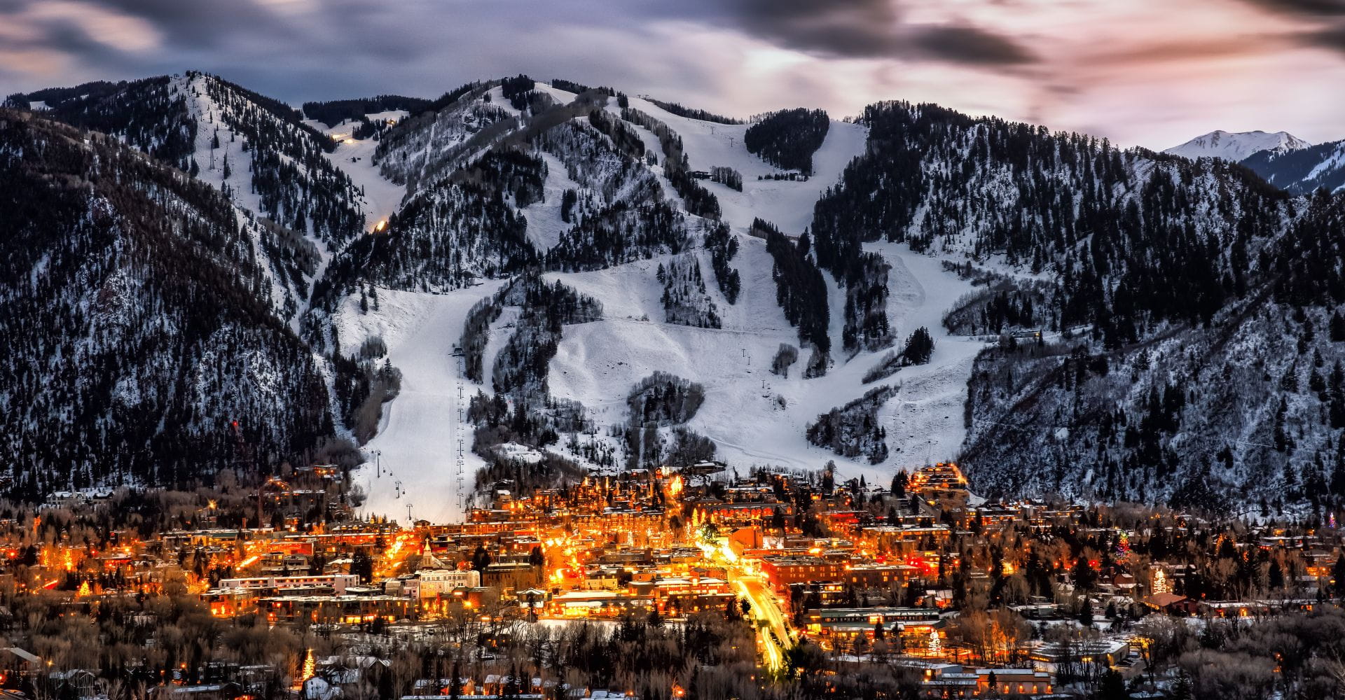 A scenic town illuminated at dusk rests beneath snowy mountains. The warmly lit streets contrast with the cool, expansive snow, creating a serene, cozy mood.