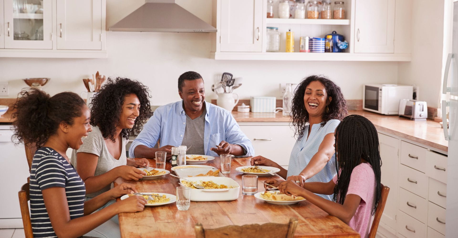 A family of five enjoying a meal together at a wooden dining table in a bright kitchen, smiling and sharing food.