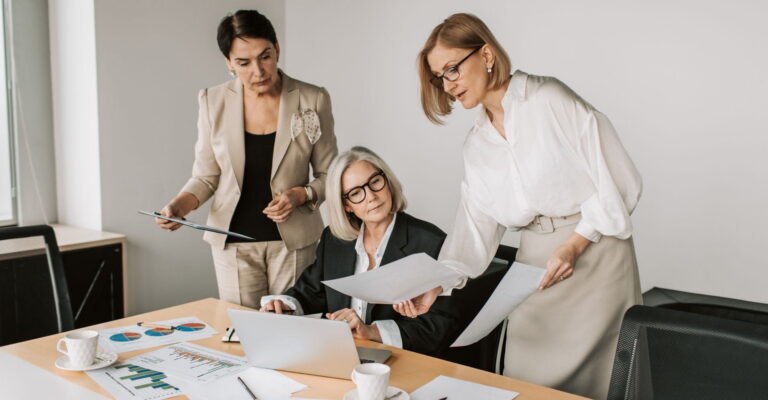 Three businesswomen discussing documents at a conference table with charts and a laptop, in a modern office setting.