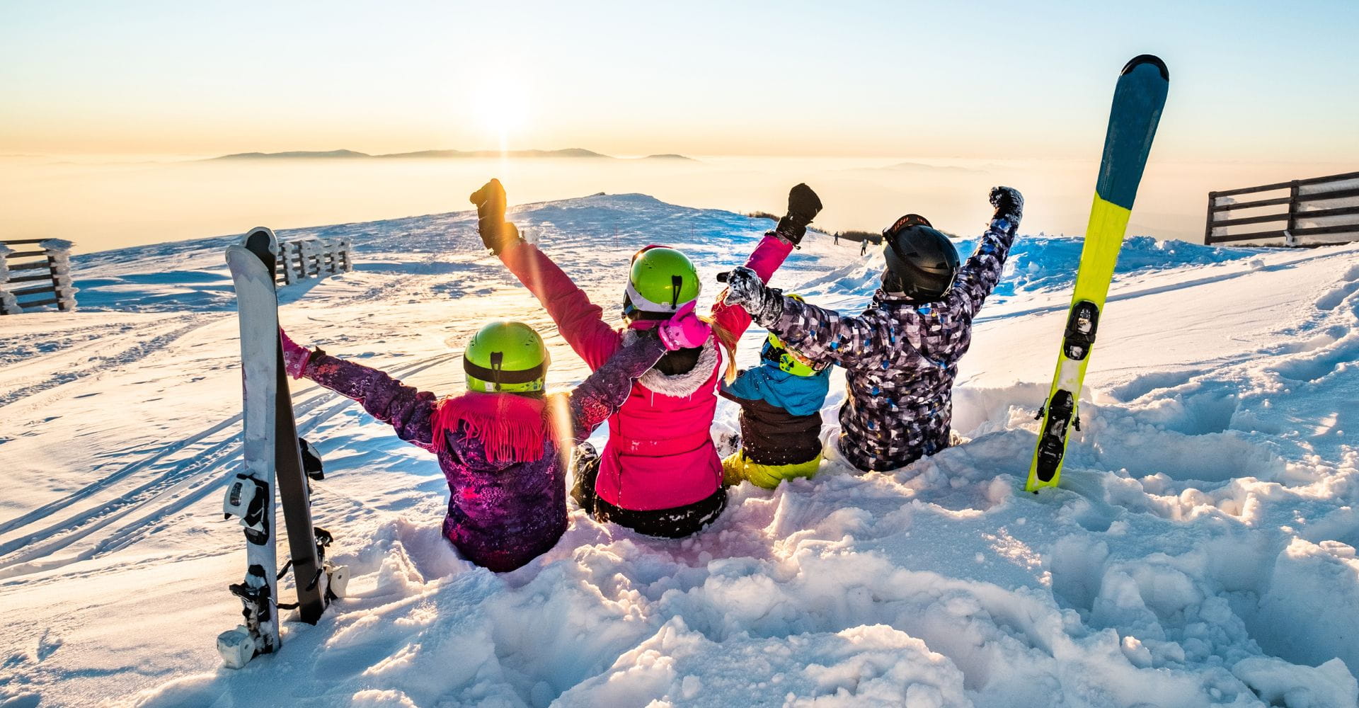 Four people in winter gear sit in the snow with raised arms, facing a snowy mountain landscape at sunset; two pairs of skis are standing upright nearby.