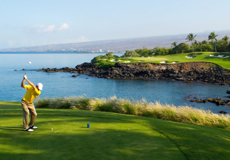 A golfer in a bright yellow shirt swings beside a blue coastal bay, with a rocky shoreline, palm trees, and a green golf course in the distance.