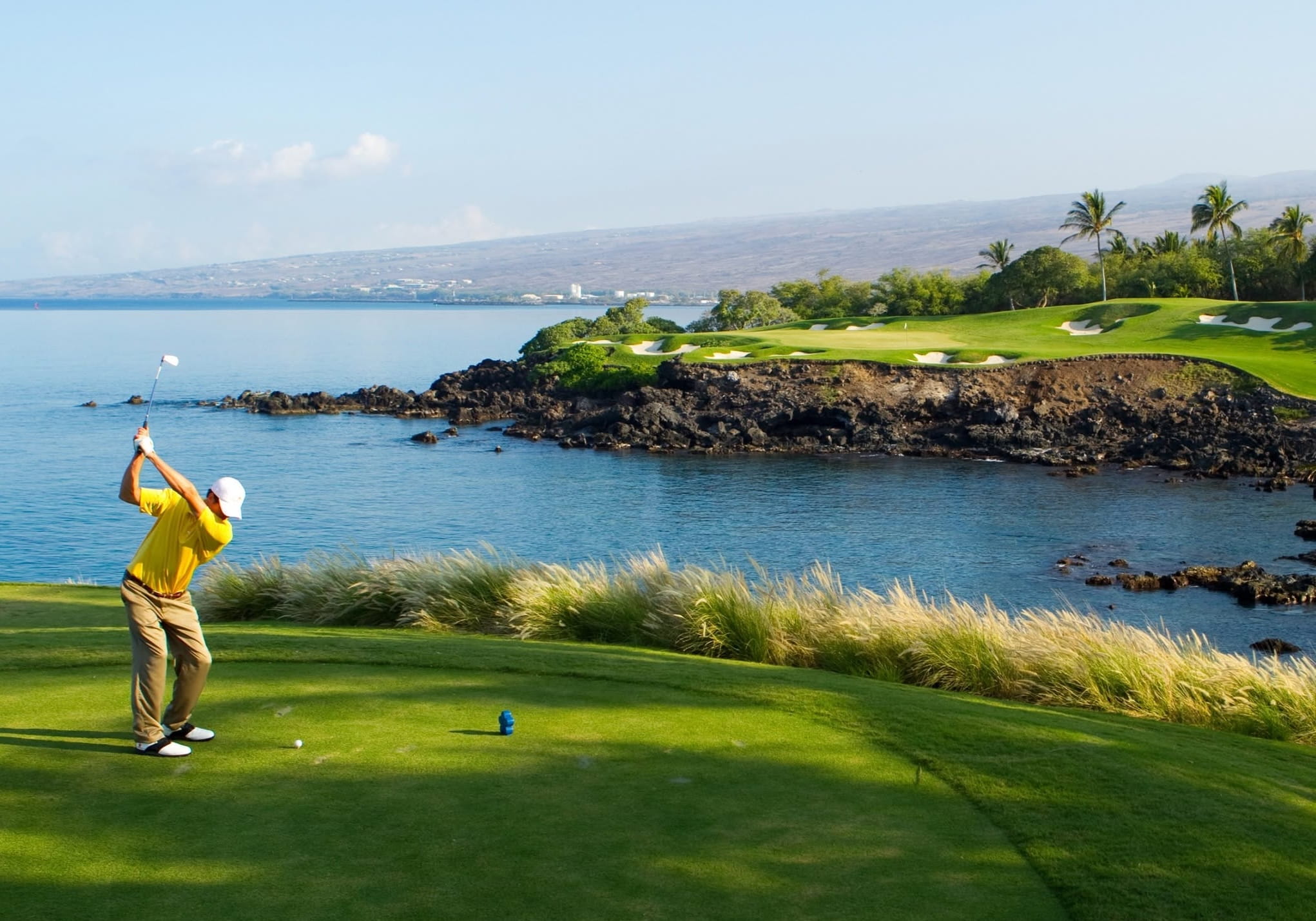 A golfer in a bright yellow shirt swings beside a blue coastal bay, with a rocky shoreline, palm trees, and a green golf course in the distance.