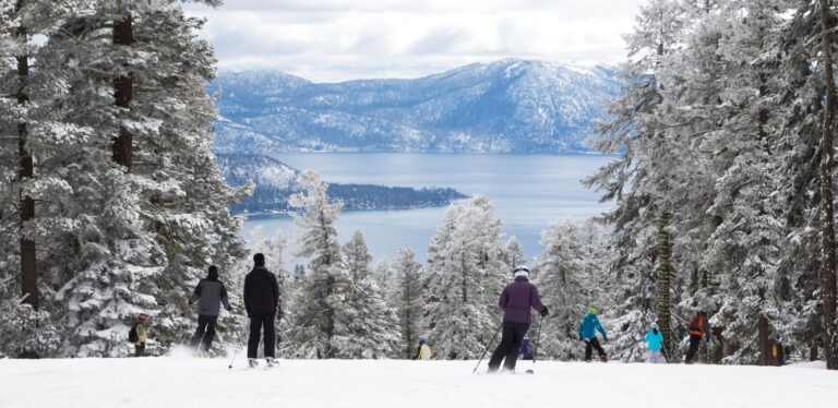 Skiers glide down a snowy slope, surrounded by trees and a serene body of water in the background.