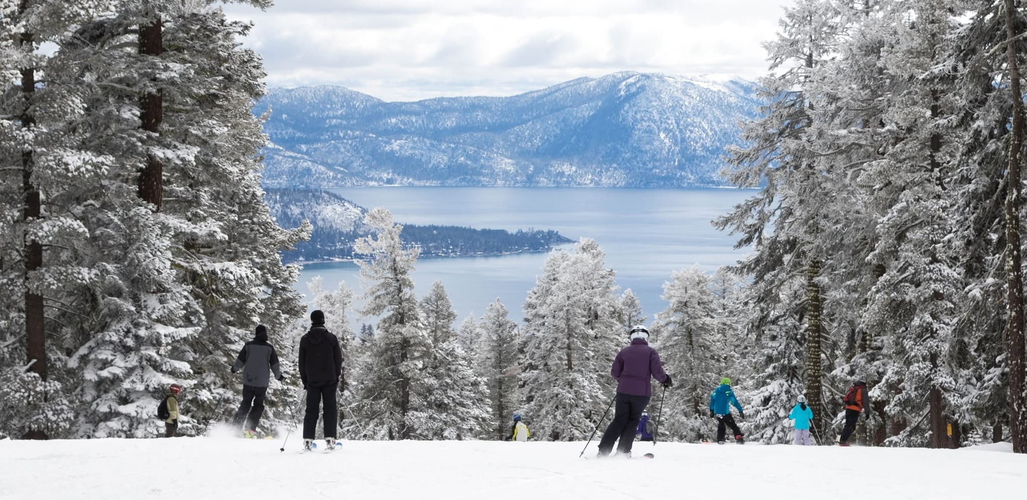Skiers glide down a snowy slope, surrounded by trees and a serene body of water in the background.